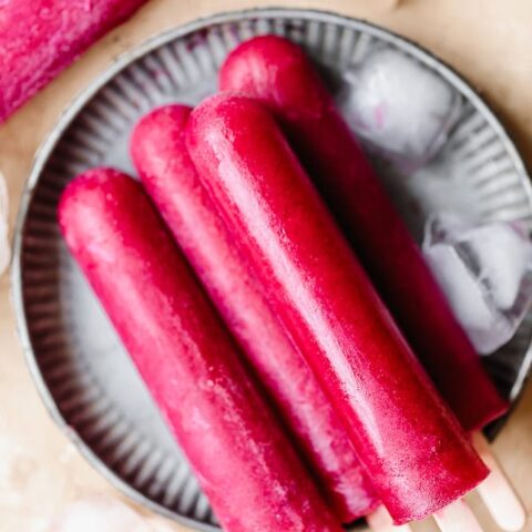 Refreshing pink fruit popsicles on a plate with ice cubes and a pink popsicle in the background.