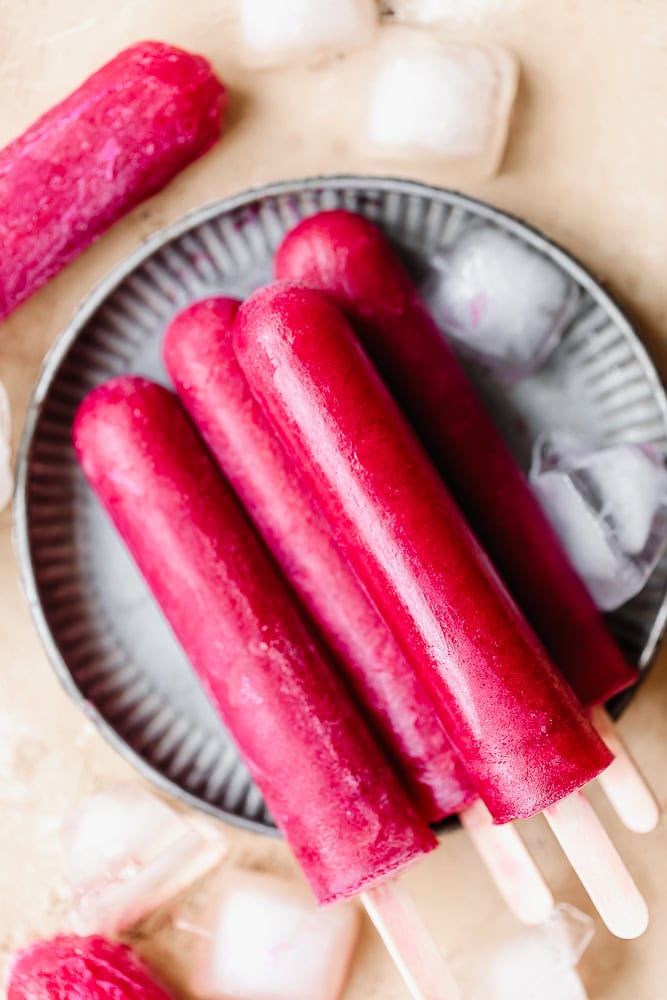 Refreshing pink fruit popsicles on a plate with ice cubes and a pink popsicle in the background.