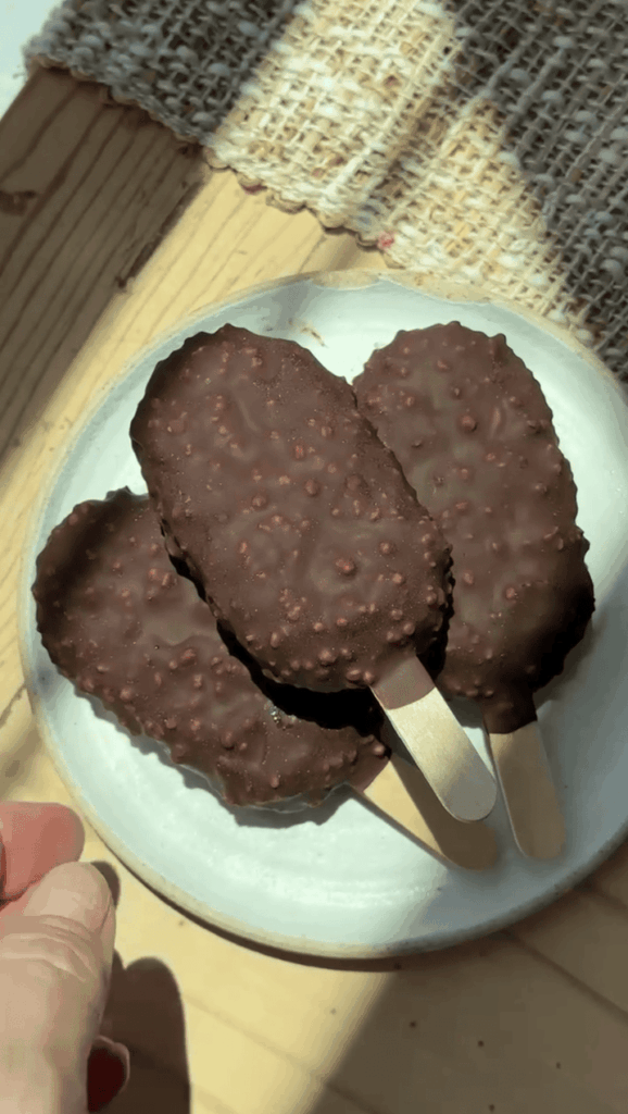 Chocolate ice cream bars with crunchy coating on a white plate in sunlight.